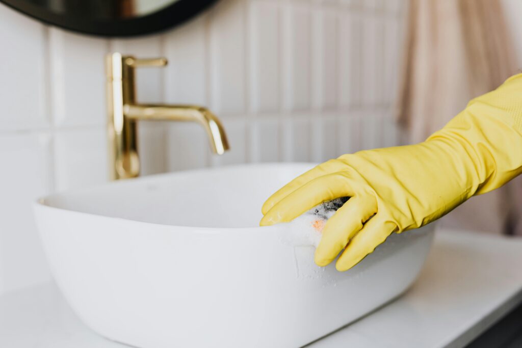 Crop faceless person in yellow latex protective glove cleaning surface of white stylish sink in contemporary bathroom interior