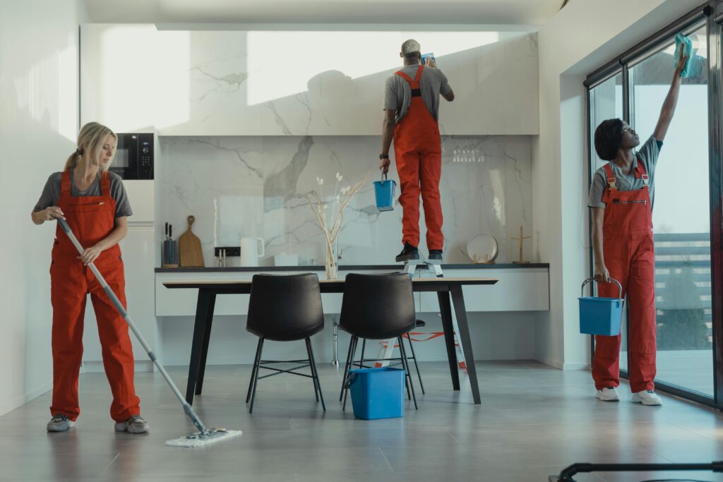 Group of cleaners in red uniforms mopping and wiping glass in a modern dining room.