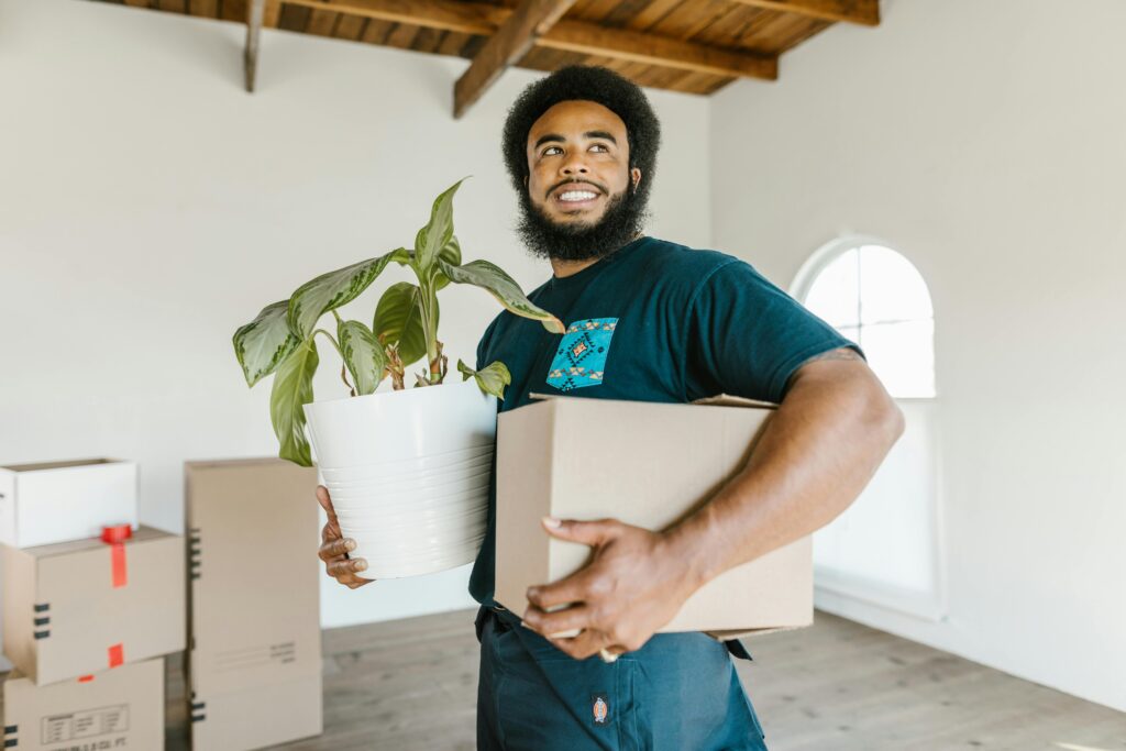 Smiling man holding a moving box and potted plant in a bright new home.